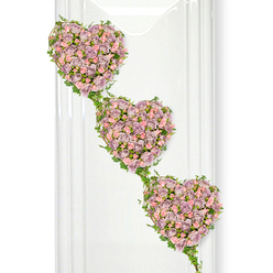 Three romantic, heart-shaped floral tributes tumble gently down a pristine white backdrop, forming a graceful diagonal line that suggests an unbroken thread of love and memory. Each heart is densely composed of miniature roses in soft dusty lavender, muted mauve, and delicate pastel pink, the colours blending like watercolour on silk. The roses are tightly clustered so that hardly any space can be seen between the blooms, creating a plush, cushion-like surface that feels almost touchably soft. Some petals curl delicately at the edges, while newer buds stand proud, adding depth and movement to the arrangement. Twining around the contours of each heart and linking them together is a garland of fresh green ivy, its small, glossy leaves catching the light and framing the roses with a natural, leafy border. The ivy trails connect the hearts in a gentle cascade, much like shared memories that travel through a family. The lighting is bright yet tender, similar to daylight streaming into a quiet room in Queensbury, not far from the calm open spaces around Queensbury Circle, giving the piece a tranquil and respectful presence. On the left, faint vertical reflections hint at a polished panel or glass, subtly suggesting a contemporary setting without drawing attention from the flowers. This triple-heart design feels especially suited to a funeral or memorial gathering, echoing themes of unity, togetherness, and enduring affection, and it would sit beautifully beside a photograph or candle during a service in the local Queensbury area.
