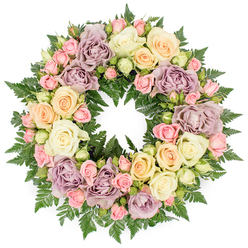 A full, circular funeral wreath is displayed against a clean white backdrop, its composition of roses and foliage forming a tranquil, elegant statement suitable for a memorial gathering in Queensbury. The wreath is densely composed, giving the impression of generous abundance, with hardly any gaps between the blooms. Wide, open roses in soft ivory and creamy white dominate the design, their layered petals gently ruffled at the edges, catching the light in subtle highlights and shadows. Around them, smaller blush pink spray roses appear in varying stages of bloom, from tightly wrapped buds to partially opened blossoms, suggesting both farewell and the continuity of life. Interwoven among these are soft peach and pale yellow roses, which introduce a gentle glow of warmth, while distinctive dusty lavender and muted mauve roses provide a vintage, contemplative note that deepens the emotional tone. The blossoms rest on a bed of rich, dark green fern fronds, whose fine, feathery texture emerges between the flowers, framing them and giving depth to the arrangement. The crisp white background keeps all the focus on the wreath itself, enhancing the clarity of each pastel shade and the fresh gloss of the foliage. The overall design feels calm and composed, the classic circular form symbolising eternity and unbroken love, making it a fitting tribute for services at local venues near Queensbury and along nearby Honeypot Lane. Seen as a whole, the wreath conveys quiet comfort, reverence, and a message of sympathy expressed in the gentle language of roses and ferns.