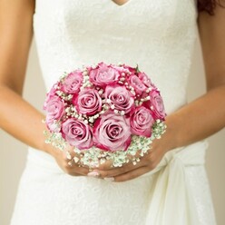 Set against the tranquil, muted backdrop of a sunlit wedding morning in Queensbury, a single, elegant bridal bouquet is highlighted with striking clarity. The round, compact cluster of roses glows with layered shades of pink, each bloom artfully transitioning from a whisper-soft blush at its center to rich, saturated magenta at its edges-a gradient that evokes painterly brushstrokes and the vibrant palettes seen in local Queensbury parks come spring. Wispy clouds of pristine white baby's breath weave through the roses, lending the arrangement an ethereal air and a sense of gentle movement. The bride's warm, subtly tanned hands, carefully manicured with a hint of glimmering nude polish, cradle the arrangement with visible care and anticipation. Her wedding dress, a sleeveless dream adorned with delicate floral lace and tailored with a flowing sash, creates a poetic echo of the bouquet's romantic textures. This photograph, captured near the leafy crescents of Honeypot Lane, encapsulates the heartfelt artistry and excitement of a couple's special day. It's a scene familiar to every local florist: the joy of crafting a bouquet that elevates a bride's moment and weaves together both tradition and the distinctive character of a Queensbury celebration.