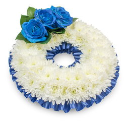 Captured with delicate attention to light and composition, this circular funeral wreath rests on a pristine white background and casts a soft shadow that enhances its modest elevation and presence. The dominant ring is composed of densely packed white chrysanthemums; their feather-soft petals overlap in a full, textured band, and the faint, creamy yellow centres peek through, bringing a gentle warmth to the composition. On the upper left an intentional cluster of three deep electric-blue roses rises from the cushion of chrysanthemums - their velvety petals unfurl with measured dignity, saturated in a calming sapphire hue. Rich, dark green foliage cradles the blue roses, adding depth and an organic outline to the focal cluster. A pleated satin ribbon in the same cobalt blue trims both the outer and inner edges of the chrysanthemum ring, the glossy folds catching light to lend structure and a touch of reverent formality. The overall effect is one of quiet sophistication: textures that invite the eye to linger, colours that speak of remembrance, and craftsmanship that reflects careful handwork. As a florist serving Queensbury, from near Queensbury station through the surrounding streets toward Kingsbury and Wembley, I often recommend this palette for memorial services and graveside gatherings; its tone is neither ostentatious nor stark, but composed - a comforting emblem of condolence. Flower Delivery Queensbury prepares such tributes with the same sensitivity and attention to freshness expected at local funeral chapels and wakes.