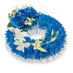An elegantly crafted sympathy wreath in deep blues and pure whites, photographed against a crisp white background to emphasize its thoughtful details. The main body consists of countless small, vivid blue chrysanthemums packed to create a plush, circular cushion that reads as both solemn and comforting. A ruffled white satin ribbon traces the inner circumference, its soft folds lending a tailored, formal border that brightens the composition. At the wreath's upper-left quadrant sits a trio of magnificent blue roses, their petals opened wide in a luxurious sapphire tone; they are haloed by tiny sprigs of gypsophila and a few dark green leaves, which add delicate contrast and a traditional touch. Flowing from the mid-left across to the lower right is a gentle cascade of white orchids - Phalaenopsis-style blooms - whose slender, porcelain-like petals bring an ethereal softness. Interspersed among them are fresh green buds, symbolic of life's continuity amid mourning. The lighting is cool and diffused, bringing out textures from velvet rose petals to the silkiness of the orchids and the matte fuzz of chrysanthemums. As a local Queensbury florist might suggest for a funeral or commemoration near Wembley or Kenton, this wreath is composed to convey profound respect and serene memory. Flower Delivery Queensbury can prepare and deliver this composed tribute with sensitivity to service times and venue requirements across the Queensbury area.