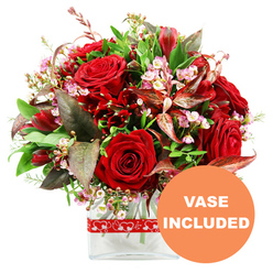 A richly detailed romantic bouquet stands out crisply against a white background, its colours and textures evoking the feeling of a carefully chosen gift delivered to a doorstep off Honeypot Lane in Queensbury. At the centre, large, velvety red roses form a full, rounded crown, each bloom at a slightly different stage of opening, from tightly wrapped buds to open cups of layered petals that seem almost touchably soft. Among these roses, smaller deep red blooms reminiscent of alstroemeria or similar multi-petalled flowers nestle closely, their trumpet-shaped or flared heads adding variety and depth to the dome-like shape. Fine sprays of tiny pale pink and white waxflower-style blossoms are scattered throughout, their minute petals and round buds creating a lace-like veil that lightens the composition and adds a gentle, romantic air. A generous frame of greenery surrounds the flowers, with broad, rich green leaves forming a sturdy backdrop and more delicate, lighter green sprigs giving a fresh, garden-like feel that brings to mind a stroll through a Queensbury park after rain. Tucked within the foliage, distinctive burgundy and russet-toned leaves with subtle veining introduce warm, autumnal notes that intensify the reds and lend the whole design a sophisticated, slightly dramatic mood. The bouquet is placed in a sleek, clear glass vase with a straight, rectangular silhouette, the transparent sides revealing the natural lines of the stems and the clarity of the water. Wrapped neatly around the vase is a bright red ribbon patterned with repeating white heart outlines, a playful yet elegant nod to romance. In the bottom right of the image, a bold orange circle carries the words "VASE INCLUDED" in clean white capital letters, signalling that this floral arrangement arrives as pictured, ready to grace a table or windowsill anywhere in Queensbury without extra preparation.