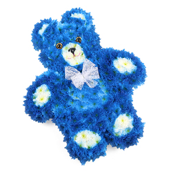 A tender floral teddy bear, carefully shaped from living blooms, lies against a crisp white background, its royal blue colour deep and comforting. The entire form is built with small, densely clustered flowers - likely chrysanthemums or similar florets - so close together that they mimic the plush pile of a beloved soft toy. The bear's round head, plump tummy, and outstretched paws are all defined by this tapestry of petals, while accents of creamy white and soft butter yellow flowers gently carve out the inner ears, the rounded muzzle, and the oval pads on each paw. These lighter tones bring a friendly warmth to the blue, adding clarity to the features without losing the overall serenity of the design. Two dark, glossy eyes and a neat black triangle nose, set above a subtle curved smile, give the bear a peaceful, almost watchful expression. Around its neck, a sheer white organza-style bow is tied with care, its ruffled edges and gentle transparency adding a note of grace. The bear appears to be slightly tilted, which lends a sense of naturalness, as though someone has just placed it with love. In Queensbury, this kind of floral tribute would feel at home as a thoughtful memorial piece delivered to a family near Honeypot Lane or Church Lane, particularly when honouring a baby boy or young child. It carries themes of innocence, remembrance, and quiet protection, crafted by a florist who understands how such tributes can gently cradle the memories of those we hold closest to our hearts.
