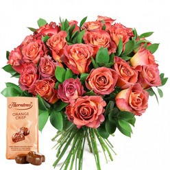 The image reveals a scene of pure gift-giving delight as experienced by a Queensbury florist-a deeply radiant, rounded bouquet of roses set against an immaculate white background, brimming with expressive energy. Each rose unfurls with painterly grace, beginning in glowing coral or soft golden peach at its core before shifting outward through subtle gradients of salmon and rich pink, their ombre petals glowing in the gentle studio light. Lush greenery, both deep and glossy, cradles the blossoms and creates a sense of fullness that is both dynamic and serene; the foliage might be eucalyptus or salal, its rich green shade amplifying the warmth and richness of the blooms themselves. To the lower left, an elegantly simple Thorntons Orange Crisp chocolate pouch sits upright, its signature logo and bold typography framed by a tempting image of two glossy milk chocolate truffles. Arranged thoughtfully in front are two real truffles, their chocolate shells gleaming as if freshly hand-wrapped, inviting both sight and taste. Crafted by a local artisan near Queensbury Circle or Roe Green Park, this bouquet-and-chocolate set is ideal for every sweet and significant celebration, whether a milestone birthday, heartfelt thank you, or a cherished anniversary. It's a composition that beckons the senses: the freshness of local roses, the crisp contrast of greenery, and the indulgent promise of chocolate. Each element is designed to evoke comfort, joy, and the warmth that comes from giving or receiving flowers in Queensbury's close-knit community.