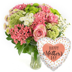 Set against a simple white backdrop, this exuberant Mother's Day arrangement feels like a burst of happiness ready to be delivered to a doorstep in Queensbury. The bouquet forms a full, rounded dome in a clear glass vase, showing off tidy green stems and sparkling water beneath a canopy of colour. Dominating the composition are generous pink roses, their petals layered in tight whorls with softly deepened edges, creating a gentle gradient from warm rose in the centre to paler pink toward the tips. Around them, clusters of coral-pink flowers with small, starry petals resemble kalanchoe, adding an almost waxy, resilient texture that contrasts beautifully with the roses' softness. Lime-green button chrysanthemums punctuate the design like bright little fireworks; their compact, cushiony blooms provide a fresh, zesty energy that instantly catches the eye. Scattered throughout are white daisy-like spray chrysanthemums, their golden centres and clean white rays lifting the palette and evoking crisp spring air in nearby Queensbury Park. Dark, glossy foliage cradles the blossoms, creating a natural frame that grounds all the vibrant tones. To the lower right, a heart-shaped foil balloon floats playfully, its light silver-white background dotted with irregular gold and peachy polka dots that shimmer as though catching afternoon light along Streatfield Road. At the centre of the balloon, a solid peach heart carries the message "HAPPY Mother's DAY" in stylish black lettering, tying the floral colours together. The overall mood is bright, grateful, and celebratory, a thoughtfully coordinated gift set that combines florist artistry with a joyful keepsake for mums across Queensbury.