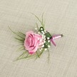 A delicately crafted pink rose boutonniere is displayed on a softly textured, light beige linen cloth, the kind of refined detail you might choose for a Queensbury wedding near the village high street. From a slightly elevated angle, the single blush-to-rose-pink bloom sits at the centre of the image, its velvety petals unfolding in gentle spirals from a paler pastel heart to deeper pink edges that curl outward with natural grace. Tiny white baby's breath clusters nestle around the base of the rose, their cloud-like blossoms adding a misty halo of softness and classic romance. Fine, grass-like strands of bright green foliage fan lightly from behind, giving the piece a sense of movement and airiness, while a single darker green leaf is neatly folded to cradle the stems, creating a tidy, architectural base. The stems themselves are bound with a narrow satin ribbon in complementary pink and lavender tones, tied into a simple, elegant knot that adds just a glimmer of sheen without overwhelming the flowers. Soft, even lighting falls across the scene, catching the gentle sheen of the ribbon and the subtle weave of the linen, while casting tender shadows that give the boutonniere a three-dimensional presence, as though it has just been set down before being pinned onto a groom's lapel. This image captures the quiet excitement of a special day in Queensbury, from prom photos in local parks to intimate ceremonies, and reflects the considered craftsmanship a local florist brings to every small but meaningful floral detail.