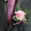 This image focuses tightly on the chest and shoulder of a well-dressed gentleman in Queensbury, ready for a milestone occasion such as a wedding, prom, or black-tie dinner. The dark charcoal suit jacket, crisply tailored, creates a strong, elegant frame for the details layered over it. Underneath, a light dress shirt with a faint gingham or check pattern peeks out at the collar and placket, offering a subtle texture that contrasts with the smooth, lustrous magenta tie. The tie falls neatly down the centre of his torso, its rich pink tone immediately drawing the eye and setting a romantic mood. Pinned precisely to the left lapel is a lovingly made boutonniere, the kind a local florist at Flower Delivery Queensbury might prepare for a ceremony at a nearby hall or a celebration overlooking one of the green spaces off the high street. A single pastel pink rose takes centre stage, its petals unfurling in perfect symmetry from a deeper rose-pink heart to softly blushed outer edges. Clusters of white baby's breath nestle around it, their cloud-like sprays lightening the arrangement and suggesting a hint of airiness. Behind these, thin, lively strands of fresh greenery spring upwards and outwards, giving the piece a natural, almost garden-picked charm while remaining polished and refined. The stems are gathered and concealed by a deep green wrapping, finished with a satin-finish ribbon in a gentle lavender-purple, tied into a small, deliberate bow. The slight sheen of the ribbon mirrors the tie, pulling all the colours together. The photograph, with its calm lighting and close framing, conveys tenderness, care, and the promise of a joyful gathering in the heart of Queensbury.