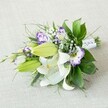 A hand-tied bouquet of white lilies and purple lisianthus rests diagonally across a softly textured light beige fabric, as if just set down on a table in a quiet Queensbury sitting room. The lilies are the undisputed stars: large Oriental blooms with creamy white petals opening wide, each petal dusted with fine reddish speckles and circling prominent yellow-orange stamens that catch the light. Beside them, several plump, chartreuse-green lily buds stretch forward, still closed but clearly full of promise, suggesting the flowers will continue to open over the coming days. Threaded between the lilies are ruffled lisianthus blossoms, their cup-shaped petals dipped in rich purple along the frilled edges and softening into white toward the centre, with tones that shift from cool lavender to deep violet depending on how the light touches them. Tiny clouds of baby's breath float in and out of view, their miniature white flowers adding a misty, romantic layer that softens the overall design. A wealth of greenery grounds the bouquet: broad, glossy dark green leaves provide a strong, structured base, while finer, feathery fronds and a few spikier green stems create movement, almost like a breeze moving through the arrangement. The stems are neatly gathered and wrapped in a crisp white satin ribbon, delicately finished with tiny pearl-like beads along the narrow end, giving the piece a bridal, ceremonial feel. The light is gentle and even, with no harsh shadows, so every texture-from the satin-smooth lily petals to the slightly nubby fabric beneath-feels calm and serene, ideal for a wedding bouquet or elegant gift delivered to a home near Queensbury Park.