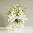 An exquisitely balanced wedding bouquet is captured in clear, even studio light, evoking the quiet elegance of an intimate ceremony in Queensbury. Several pristine white lilies open wide at the center, each bloom like a graceful star, their smooth petals curving outward to reveal delicately speckled throats and warm yellow-orange stamens. Around them, white lisianthus blossoms and buds mingle, some fully ruffled with soft lavender-tinted edges, others still closed in pale green pods that promise to unfurl in the days ahead. Fine sprigs of dark green foliage, lacy white filler flowers, and small clusters of rich blue-purple blossoms thread through the design, adding depth and a gentle contrast that keeps the arrangement from feeling overly stark. The flowers are gathered into a round, sculptural vase formed from crisscrossing white strands, almost like a contemporary basket, its open structure allowing glimpses of shadow and light. Resting on a faultless white surface, the vase is flanked by two natural woven spheres in a light rattan tone, which mirror the textural interest of the container and bring a subtle, earthy touch. Behind, a vertically textured beige background lends a soothing, fabric-like softness, reminiscent of a carefully dressed reception space along Church Lane or near Queensbury's quiet residential streets. This bouquet feels ideal for wedding collections, vow renewals, or a refined thank-you, the kind of arrangement that Flower Delivery Queensbury might send same day to celebrate love, offer comfort, or grace a modern home with a touch of tranquil floral artistry.