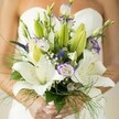This Queensbury bridal bouquet whispers of elegance and hope, brought to life in a carefully arranged armful of blossoms. Creamy Oriental lilies open generously, their luminous petals and sunlit yellow stamens drawing the eye while their delicate fragrance lingers softly-much like blossoms seen in the gardens of Canons Park. Intertwined lisianthus flowers, edged in watercolour lavender, dance lightly amidst the pure white blooms, hinting at playfulness and the promise carried by wedding mornings in Queensbury. Sprigs of deep purple Veronica and airy clouds of baby's breath provide movement and dimension, their textures adding both gentle wildness and sophisticated harmony. Vibrant green foliage, both broad and wispy, spills over the sides, cradling the bouquet in a cascade of natural abundance. At the base, the bride's manicured hands, elegant and light, reflect the grace and anticipation of the moment, as her white gown blurs softly behind, allowing the bouquet to steal the scene. Each flower, every leaf and tendril, is arranged to convey a sense of joyful anticipation-a perfect touch for a day when love and artistry unite in Queensbury.