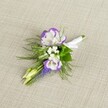 An intricately crafted wedding boutonniere lies against a softly textured, light beige linen background, its neutral tones allowing every detail of the flowers to stand out with quiet sophistication. At the heart of the design, two open lisianthus blooms display pristine white petals delicately ruffled and edged in a deep, regal purple, giving the impression of hand-painted borders that draw the eye inward. Tucked around them are airy flecks of tiny white filler flowers, reminiscent of baby's breath, adding a cloud-like softness and echoing the freshness of early morning walks across Queensbury's parks. A slender vertical spike of tightly clustered, bell-shaped muscari in rich purple-blue rises from the side, its beaded texture introducing depth and a touch of whimsy. Fine, feathery greenery, similar to asparagus fern, fans out in gentle sprays, its fresh green hue and delicate fronds suggesting the light movement of a breeze along a quiet street near Queensbury Circle. Two pale green, unopened buds nestle among the foliage, hinting at blossoms still to come and lending a sense of promise to the arrangement. The stems are carefully bound together and wrapped in a crisp white satin ribbon, smoothed neatly and finished with a small, refined bow that signals it is ready to be pinned to a lapel. The lighting is soft and even, making the colours appear true and calm, as though the piece has just been placed down by a florist preparing for a ceremony in a nearby Queensbury venue, ideal for a groom, groomsman, or prom guest who prefers understated elegance and gentle romance in their flowers.