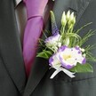 A close-up wedding portrait focuses on the upper torso of a suited gentleman, showing only his dark charcoal grey jacket, light grey check shirt, and rich magenta silk tie, with the camera lingering tenderly on an exquisite lisianthus buttonhole. The tailored suit fabric appears softly textured, lit by gentle indoor light that picks up the faint pattern in the shirt and the luminous sheen of the tie, giving the scene a polished yet relaxed feel, like a special day unfolding in Queensbury's town centre. Pinned carefully to the left lapel, the boutonniere features two open lisianthus blossoms, their white petals edged in deep, painterly purple ruffles that curl slightly at the tips, and bright yellow centres that glow against the darker suit. Around them, several slim, unopened buds reach upward, promising future blooms, while a slender vertical spire of small, indigo-purple veronica florets runs alongside, adding height and a touch of wild garden character reminiscent of flowers growing near Queensbury Park. Delicate sprays of tiny white filler flowers, much like baby's breath, weave through the design, softening the lines and creating a cloud-light halo around the feature blooms. Fresh greenery in differing textures-feathery fronds, smooth emerald leaves, and a few artistic loops of fine green grass or wire-cradles the flowers and frames the tie's bold colour. At the base, a neat white satin bow holds everything securely, its subtle shine echoing the shirt's crispness. The overall impression is one of quiet, joyful anticipation: a groom or groomsman dressed and ready, wearing a small piece of floristry art close to his heart, as if he had just stepped out from a local Queensbury florist's studio on his way to say I do.