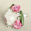 Viewed from a slightly elevated angle against a pale, subtly woven fabric background, this bouquet appears as though it has just been set down on a dining table in a Queensbury home, ready for a small family celebration. At its centre sits a plush creamy white peony, the bloom generously sized and layered with countless petals that curl and fold into one another like soft silk ruffles, giving it a luxuriously full, cushiony presence. Framing this central flower are two pink roses, one rising just above and one resting just beneath, their petals spiralling out from tight, well-formed cores. The colour gently deepens from soft pink in the middle to richer magenta tones at the edges, adding drama without losing the bouquet's refined charm. Delicate clusters of white baby's breath are woven throughout, forming a fine mist of tiny blossoms that lend the arrangement an ethereal, almost bridal veil effect, reminiscent of wedding photos taken in local parks such as Queensbury Park or Eton Grove. Around the base, glossy dark green leaves curve inward, their broad surfaces catching the light and grounding the lighter tones with a strong verdant frame. The stems are expertly gathered and wrapped in impeccable white satin ribbon, suggesting a hand-tied design meant to be held comfortably during a wedding or formal event. The light falling across the scene is soft and even, revealing every subtle texture-from the velvety petals to the slightly crinkled fabric beneath-creating an atmosphere of romance, grace, and thoughtful craftsmanship that feels at home in the quiet streets of Queensbury.