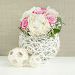 A lush, rounded bouquet of ivory peonies and vivid fuchsia-pink roses sits proudly in a white, basket-like vase, the whole scene bathed in soft, natural light that feels reminiscent of a peaceful Queensbury afternoon. The peonies are plush and full, their creamy petals cupping in gentle ruffles that suggest a soft, powdery fragrance, while the roses bring a more saturated burst of pink, each spiral of petals hinting at romance and quiet admiration. Wisps of baby's breath weave between these statement blooms, their tiny, snow-white florets forming a fine, cloud-like veil that gives the arrangement a dreamy, bridal quality. Encircling the flowers, rich green leaves fan out in a smooth, polished ring, their surfaces catching the light like newly washed foliage after rain in one of Queensbury's leafy side streets. The vase itself is a striking feature: a rounded, open lattice of slender white strands, reminiscent of handwoven rattan, giving the impression that the bouquet is floating in a delicate, sculptural cradle. Resting on the crisp white tabletop are two decorative woven spheres in a pale straw tone, one slightly larger than the other, echoing the texture of the vase and adding balance to the composition. The neutral beige background functions much like a freshly painted wall in a modern Queensbury home, allowing the colours and forms of the arrangement to take center stage. Crafted with the care you'd expect from a local boutique florist, this design would suit a wedding breakfast near Queensbury Station, an engagement dinner at home, or any occasion where grace, tenderness, and contemporary style are called for.