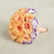 Captured from a slightly raised viewpoint on a neutral, textured beige fabric that resembles natural burlap or linen, this elegant bridal bouquet is a study in warm colour and delicate contrasts. The composition centres on a compact, dome-shaped grouping of peach and soft orange roses whose tightly layered petals create a sumptuous, velvety mass. Interspersed among them, white lisianthus edged in a striking violet introduce a cool, unexpected punctuation that brightens and deepens the arrangement without overpowering the rosy warmth. Sprays of baby's breath (gypsophila) float between the larger blooms like soft confetti, lending an airy, whimsical quality to the otherwise richly textured cluster. The bouquet's handle is wrapped in a light peach satin ribbon whose gentle sheen echoes the roses' luster; around it coils a slim strand of cream pearls, a small but luxurious touch that suggests heirloom charm and thoughtful detail. The directional light softens shadows and accentuates the ribbons of colour in each petal, while the tactile contrast-silky petals, airy gypsophila, smooth ribbon and cool pearls-makes the bouquet feel as inviting to hold as it is to look at. This floral piece reads as an emblem of commitment and celebration: ideal for a Queensbury wedding, a vow renewal at a local parish, or an intimate anniversary dinner near Fryent Country Park. The overall effect is romantic, refined and quietly joyous, evoking the scent of fresh roses and the hush of a moment worth remembering.