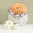 A graceful floral arrangement stands as the focal point on a bright white surface, its refined palette and textures recalling a carefully styled table at a Queensbury celebration. The centerpiece is built around a rounded dome of roses in gentle peach and apricot shades, the blooms tightly clustered so that their petals form a smooth, seamless curve, like a soft floral sculpture. Each rose appears lush and freshly opened, with the kind of velvety fullness that feels perfect for weddings or romantic occasions. Framing the lower edge of this rose sphere is a circle of white lisianthus, their ruffled petals delicately outlined with a band of rich purple that introduces a more dramatic note while still harmonising with the softer tones above. Fine sprigs of white baby's breath tuck in between, their tiny blossoms catching the light and adding a whisper of texture. The entire arrangement is nestled into a wide, bulbous container made of white woven strands, likely rattan or wicker, that criss-cross to form an open, airy lattice. This sculptural base feels both natural and contemporary, complementing the flowers without competing with them. On the tabletop in front, two decorative spheres crafted from thin, pale wooden strips sit side by side, one noticeably larger than the other, echoing the woven motif of the vase and gently extending the visual story. Behind the display, a softly textured beige backdrop provides warmth and subtle depth, reminiscent of fabric draped along a wall or behind a reception table. Together, these elements create a scene of quiet sophistication and romantic charm, ideal for imagining same-day floral delivery to Queensbury homes and intimate venues.