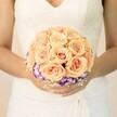 A softly lit, chest-level photograph shows a bride standing still, her hands gently cupping a perfectly round bridal bouquet in front of a classic ivory gown. The bodice of her dress is trimmed with delicate lace and a modest V-neckline, while the skirt falls in smooth, graceful folds that blur into a pale, almost cloud-like background. At the centre of the image is the bouquet itself: a lush dome of tightly clustered roses in shades of peach, apricot, and blush pink, each bloom spiralling inward with velvety, layered petals. The warm tones of the roses feel like the first light over Queensbury Park on a summer morning, soft yet radiant. Just beneath this rose canopy, a neat collar of ruffled blooms in deep royal purple with white, frilled edges-reminiscent of lisianthus-forms a striking ring, giving the bouquet depth and a touch of drama. Tiny sprigs of airy white filler flowers, much like baby's breath, peek through around the base, adding a gentle, lacy texture that echoes the gown's lace. The bride's nails are neatly manicured in a pale, neutral shade, with a subtle shimmer on one thumb that catches the light as if she has just stepped out from a registrar in central Queensbury. The overall mood is one of poised anticipation and romance, a moment held just before walking down the aisle, and the bouquet appears freshly arranged by a careful local florist, ready to be carried into a ceremony in a nearby church or intimate venue off Kingsbury Road.