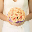 This arrangement reads like a gentle poem: a symphony of apricot roses gathered into a symmetrical cluster, each blossom unfurling with creamy, sunlit warmth. Encircling the roses is a ring of purple-tipped lisianthus whose cool, lilac-hued edges set off the peach tones like twilight framing a soft sunrise. Fine, airy filler-small white sprays that might be baby's breath-creates a delicate lattice between the main blooms, adding weightless texture and a breath of freshness to the composition. The lighting is soft and diffused, making petals appear almost translucent at their thinnest points and casting a warm glow across the bouquet's rounded silhouette. I imagine this in a bridal setting in Queensbury, carried down a quiet high street or posed for photos by the green; it complements lace and ivory fabrics and reads elegantly across black-and-white or colour images. The scent is understated and comforting: a clean, sweet rose note layered with the green, slightly herbal tones of the foliage and fillers. Crafted with precise placement and an eye for balance, the bouquet feels both traditional and modern-lush yet refined-designed to sit easily in the bride's hands while drawing the eye in photographs and memory. Its emotional tone is serene and celebratory, a small ceremony of textures and colours that speaks of new beginnings and quiet joy.