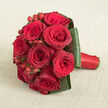 At the heart of this photograph lies an intimate posy of deep red roses, so densely gathered that they form a near-perfect sphere, cradled on a warm beige fabric that resembles finely woven linen. Each rose is at its peak, the velvety petals overlapping in intricate spirals, their hues shifting subtly from rich crimson to darker burgundy towards the shadowed centres, giving the bouquet a sense of depth and quiet intensity. Threaded between the blooms are clusters of bright red hypericum berries, their smooth, polished surfaces catching tiny highlights of light and adding a lively, almost jewel-like detail among the flowers. The bouquet is encircled by a collar of broad dark green leaves, each one carefully folded and arranged to create a structured edge, like a tailored frame that holds the roses in place. The foliage feels cool and sleek, a contrast to the plush softness of the petals and the gentle grain of the cloth beneath. Along one side, the stems are bound tightly with a bold red satin ribbon, wrapped in precise, even bands that give the handle an elegant, finished look. The overall atmosphere is romantic and composed, reminiscent of a bridal bouquet being checked for perfection in a Queensbury studio just before travelling to a ceremony near The Broadway. It is easy to imagine the faint scent of the roses, the weight of the bouquet in the hand, ready for same-day delivery across Queensbury's quiet residential streets to mark a proposal, a milestone anniversary, or a heartfelt declaration of love.