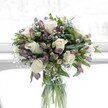 A graceful glass cylinder vase holds a full, rounded bouquet of creamy white roses, soft lavender alstroemeria, and airy white baby's breath, illuminated by gentle daylight that appears to stream from a nearby window. Each rose is in perfect bloom, its velvety ivory petals opening in layered spirals that suggest romance and quiet celebration. Around them, clusters of dusky purple alstroemeria-some still in tight buds, others just beginning to unfurl-add a cool, lilac tone, their slender petals brushed with deeper veining and held by fresh green sepals. Delicate sprays of gypsophila weave between the larger flowers like tiny clouds, creating a misty, weightless texture that lightens the entire arrangement. Lush foliage in rich, glossy greens frames the bouquet, with broader leaves at the base and finer stems reaching upward, adding height, depth, and a sense of natural movement. Through the clear vase, you can see the neatly trimmed green stems standing in fresh water, aligned with the care of an experienced florist, giving the design a clean, contemporary profile. The background is softly blurred and pale, likely a white wall or bright Queensbury window, which enhances the calm, uplifting mood without drawing attention away from the flowers. This arrangement feels serene yet hopeful, like a quiet pause in the middle of a busy day on Queensbury's high street-a bouquet suited to sending comfort, celebrating a gentle milestone, or simply bringing a breath of fresh elegance into a living room overlooking the local park. The overall impression is pure, tender, and thoughtfully crafted, as if arranged for someone who appreciates subtle beauty and classic blooms.