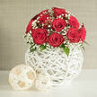 A lush dome of red roses rises from a sculptural white sphere, the whole design captured in tranquil light as though arranged for a romantic evening at home in Queensbury. Around twelve roses, all a rich, velvety crimson, are clustered closely together so that each bloom supports the next, forming a rounded crown of colour. Their petals are smooth and full, opening in graceful spirals that suggest both passion and refinement. Between the roses, sprays of white baby's breath float like tiny stars, adding a lacy edge that softens the intensity of the red and introduces a dreamy, almost ethereal quality. Discreet flashes of dark green leaves peek from beneath the flowers, offering just enough foliage to enhance the contrast without breaking the harmony of the design. The bouquet is seated in a round, pure white vase crafted from interlaced strands resembling wicker, its airy latticework giving the illusion that the flowers are almost weightless. Two lighter, cream-coloured decorative balls made of similar woven material rest to one side on a pristine white tabletop, echoing the texture of the vase and adding a sense of curated home décor. Behind, a neutral wall in soft beige or pale grey provides a quiet backdrop, like the calm interiors of a Queensbury townhouse tucked off Streatfield Road, allowing the roses to take full visual focus. Gentle, even lighting caresses each surface, revealing the interplay of smooth petals, fine gypsophila clusters, and crisp woven lines. The scene feels intimate and celebratory, suited to wedding celebrations, engagements, or special deliveries across Queensbury when you want flowers to speak in the language of timeless romance.