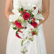 In this intimate photograph, a bride stands quietly with her hands resting at her waist, holding a cascading bouquet that seems to flow like a small waterfall of flowers over the front of her white gown. The dress itself is finely detailed, with subtle lace and embroidery that catch in the soft light, a sight reminiscent of wedding mornings in Queensbury, perhaps near the tree-lined streets around the station. At the centre of the composition, velvety red roses glow with a rich, wine-deep colour, each bloom tightly layered and full, suggesting devotion and strength. Between them, generous white lilies open wide, their broad petals both crisp and soft, with green stamens drawing the eye toward their fragrant hearts. From the lower edge of the bouquet, elegant white Phalaenopsis orchids trail downwards, their arching stems lined with open blossoms and buds, forming a gentle, rhythmic cascade that mirrors the fluid teardrop silhouette. Clouds of baby's breath weave through the arrangement, scattering tiny white stars of bloom that make the larger flowers appear to float. Small, glossy red berries, likely hypericum, dot the greens like festive jewels, adding bright points of texture and colour. Layers of foliage - from slender, fern-like fronds to broader, deeper green leaves - frame the bouquet, giving it structure and a natural, garden-grown feel. The softly blurred beige background removes all distraction, ensuring the eye remains on the interplay of red, white, and green. It is an image that perfectly conveys the romance and care behind a carefully crafted wedding bouquet, the kind a local Queensbury florist would design for a ceremony filled with emotion and quiet joy.
