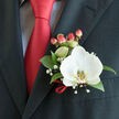 This artfully arranged boutonniere, photographed in close-up against a charcoal suit jacket, reads as a study in contrast and craftsmanship. A single white Phalaenopsis orchid takes centre stage, its smooth petals wide and creamy, the yellow-gold patterned lip drawing the eye like a tiny sunlit detail. Complementing the orchid are glossy, deep-green leaves that provide a grounding backdrop, and a few stems of tiny white gypsophila that float like distant stars, softening the silhouette. A cluster of coral-tinged hypericum berries offers a tactile pop of colour that softly mirrors the bold red satin tie beside it; a neat knot and the hint of a light-coloured patterned shirt complete the tailored picture. A slim twist of red ribbon is looped around the base of the floral pin, a subtle flourish that unites floral design with menswear in a way we often prepare for weddings, anniversaries, and black-tie gatherings across Queensbury and nearby Kingsbury. As a local florist, Flower Delivery Queensbury composes details like this to stand up to close inspection - every leaf trimmed, every stem wired and secured so the boutonniere sits flat against the lapel without twisting. The lighting here is soft and ambient, lending a gentle sheen to the orchid and a cool, tactile sensation to the foliage. The overall mood is poised and celebratory, a small emblem of care and ritual that will last through photographs, vows, and after-dinner toasts throughout the neighbourhood.