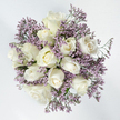 Captured from a bird's-eye view, this round bouquet rests on a bright white background, its soft colours and shapes creating the same quiet calm you might feel wandering through the greener corners of Queensbury. At the centre, a cluster of about twelve white roses forms a perfect circle, each bloom in its own serene stage of opening. Some are small, tightly held buds, their outer petals smooth and closely wrapped, while others are slowly unfolding, exposing layers of creamy, velvety petals curling outward in relaxed spirals. A few blossoms sit fully open, revealing delicate, concentric whorls with hints of warm ivory and the faintest suggestion of pale yellow in their depths, where light seems to settle. Encircling and filling the spaces between these roses is a soft cloud of lavender limonium, its myriad tiny flowers gathered along slender stems. The limonium's fine, misty texture contrasts with the solid silkiness of the roses, creating a sense of depth and gentle movement, as though the darker lilac hues are quietly cradling the purity of the white blooms. Subtle strokes of green stems and minimal foliage appear here and there, just enough to remind you of the bouquet's natural origins without interrupting the elegant colour harmony. The overall effect is tranquil, almost meditative, with even lighting and no distractions. In Queensbury, arrangements like this are often chosen as sympathy flowers for local services and home deliveries-a thoughtful, understated tribute designed to bring solace, dignity, and a soft touch of hope during times of loss.