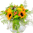 This vibrant display is a true vision of sunshine and joy, perfect for bringing the warmth of Queensbury's summer parks into any home. Dominant sunflowers, their golden faces glowing with life, stand at the heart of the arrangement, each petal radiating outward like beams from the midday sun. Their deep, velvety centres draw the eye, setting a cheerful tone that harmonises with flurries of yellow filler blooms - reminiscent of wildflowers scattered along Kingsbury Road. Small coral-orange flowers, hidden among the lush greenery, lend a playful burst of colour, their delicate forms echoing the gentle excitement of a festive neighbourhood gathering. Fronds of fluffy dark green fern arch outwards, each leaf creating elegant lines that soften the bouquet's silhouette, while clusters of silver-blue eucalyptus and zesty lime-green foliage add cool undertones and vibrant contrast. The clear, round vase rests quietly beneath, revealing the natural tangle of fresh stems immersed in sparkling water - a subtle reminder of the arrangement's garden-fresh origins. Ideal for celebrating birthdays, anniversaries, or simply brightening a loved one's day, this bouquet encapsulates the heart of Queensbury: lively, welcoming, and full of color. Its composition is both refined and free-spirited, expertly crafted to deliver happiness and warmth into the spaces and moments that matter most.
