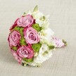 A luxurious bridal-style bouquet is photographed from a slightly raised angle, resting on a softly textured light beige fabric that feels like the calm backdrop of a Queensbury townhouse interior. At the centre of the design, several large roses open fully in a romantic swirl of lavender-pink, their velvety petals deepening to a richer, almost fuchsia edge that gives each bloom a gentle two-tone glow. Between the roses, bright chartreuse green button chrysanthemums cluster in rounded tufts, their tiny petals forming tight domes that add playful texture and a vivid splash of freshness. Threaded throughout the bouquet are airy white flowers, likely lisianthus, with delicate, layered petals and faintly greenish-yellow centres that catch the light, suggesting purity and softness, like clouds drifting over Queensbury Park. A few closed buds in fresh green hint at new blossoms still to come, reinforcing the sense of just-cut garden vitality. All the stems are neatly pulled together into a compact, rounded shape, forming a tidy handle wrapped in smooth pink satin ribbon that gleams gently, as if lit by soft afternoon light through a Bayton Road window. Spiralling around the ribbon, a strand of tiny white pearls creates an elegant, heirloom feel, evoking classic wedding traditions while still feeling modern and chic. The combination of pink roses, bright green chrysanthemums, and white lisianthus suggests romance, joy, and celebration, making this design ideal for a Queensbury wedding, an engagement celebration, or as inspiration for contemporary bouquet trends that mix classic charm with fresh, vivid colour. Even without scent, you can almost imagine a gentle floral fragrance, ready to be carried down an aisle or presented as a heartfelt gift.