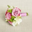 A small, elegant bridal-style bouquet lies horizontally on a pale tan, textured linen surface, the soft weave of the fabric echoing the relaxed charm of a Queensbury townhouse table. At the centre of the posy, three lush pink roses are fully open yet still deliciously layered, their petals moving from a gentle blush at the heart to deeper magenta-pink at the ruffled outer edges, like a delicate ombré sunset over nearby Queensbury Park. Nestled closely around them are neat spheres of lime green button chrysanthemums, their tiny, tightly packed petals forming playful pom-pom shapes that contrast beautifully with the smooth, velvety rose petals. Threaded between these focal blooms are sprays of creamy white and ivory flowers, possibly lisianthus or freesia, their soft, papery petals and subtle curves bringing a feeling of airiness and romance, as if ready to be carried down the aisle at a local church off Kenton Road. Tiny star-shaped white blossoms with sunny yellow centres add intricate detail, catching the gentle, even light that falls across the scene without harsh shadow. The short stems are carefully gathered and bound at one end with a vivid fuchsia satin ribbon, forming a polished handle that glows against the neutral fabric. Over the ribbon, a fine strand of white faux pearls is wrapped in a loose spiral, crossing diagonally like delicate jewellery on a bride's wrist, before the neat green stem ends emerge, freshly trimmed. The overall look is fresh, refined, and quietly joyful, a compact bouquet that would suit a bridesmaid, a young bride, or a special celebration dinner at home in Queensbury, its colours and textures radiating intimate celebration and thoughtful craftsmanship.