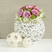 A serene, studio-style photograph showcases a refined floral centerpiece that could easily grace a wedding table at a small Queensbury reception or a stylish dinner near Mollison Way. The arrangement is composed as a perfectly rounded dome, overflowing with roses in shades of blush pink, rose pink, and light lavender. Each bloom appears carefully selected, petals unfolding in tight spirals and soft curls, with some edges deepening into a richer pink that suggests maturity and fragrance. Interspersed among the roses are small, vivid green button chrysanthemums, their compact, spherical heads creating playful dots of colour and texture that enliven the pastel palette. Around the lower edge of the bouquet, subtle clusters of tiny white blossoms weave in and out, adding a halo of softness and enhancing the sense of fullness without overwhelming the main flowers. The bouquet sits in a distinctive white spherical vase, crafted from thin, rattan-like branches intricately woven into an open, airy lattice, reminiscent of handcrafted decor found in contemporary Queensbury homes. The vase rests on a spotless white tabletop, where two decorative wicker spheres in pale natural tones sit to the left, one larger and one smaller, mirroring the woven pattern of the container and adding visual rhythm. The background is a softly blurred panel of light beige fabric with a gentle, tactile texture, likely linen or burlap, which bathes the scene in warmth and allows the pinks, greens, and whites to stand out clearly. The lighting is soft and even, revealing every curve of the petals and every strand of wicker, making this image perfectly suited to illustrate wedding centerpieces, elegant home styling, or bespoke floral designs available for local delivery throughout Queensbury.