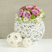 A refined floral centerpiece sits serenely on a smooth, reflective white surface, as if prepared for a wedding reception in a chic Queensbury townhouse. The main bouquet forms a rounded dome of flowers, dominated by lush pastel pink roses whose velvety petals curl outward in generous layers, some kissed with deeper pink along the edges to create depth and romance. Nestled between them are clusters of vivid chartreuse-green button chrysanthemums, their compact, button-like heads adding a lively contrast in both color and texture. At the base, an airy ring of small white blossoms and buds - perhaps lisianthus or dainty spray blooms - tumbles softly over the container, creating a gentle, cloud-like edge. These flowers are cradled in a distinctive spherical vase made from interwoven natural branches, painted crisp white and shaped into an open, basket-like lattice that feels both rustic and contemporary. The vase's structure echoes the understated architecture one might see along the quieter streets off Honeypot Lane, blending organic charm with modern design. To the left, two decorative wooden spheres in pale, natural tones rest on the tabletop, their ribbon-like strips curved and crisscrossed in an open framework that mirrors the vase's texture. The background is a warm, neutral taupe, slightly blurred so the eye stays fixed on the flowers' soft hues and intricate forms. This composition conveys calm joy and considered elegance, perfect for a Queensbury couple planning a modern wedding or for same-day delivery as a refined centerpiece to mark anniversaries, engagements, or special family gatherings.