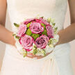This bridal bouquet rests against the intricate lace of a wedding gown, capturing a moment of stillness before the celebrations begin in Queensbury. The eye is first drawn to the lush roses at the centre, their colour a refined blend of lavender, lilac, and dusty pink that feels both contemporary and timeless. Each rose is fully formed yet not over-open, with layers of petals curling softly outward like small, contented sighs. Tucked between them, lime-green button chrysanthemums peek out, their rounded, textured blooms adding a playful spark of colour and a hint of modern design. Around the outer edge, creamy white flowers and slender buds create a soft halo, almost cloud-like, gently framing the richer tones while keeping the overall palette light and bridal.

The bouquet is round, hand-tied, and neatly compact, sitting snugly in the bride's hands as she prepares to walk down the aisle, perhaps at a nearby church or venue off Honeypot Lane. The stems are trimmed evenly, likely wrapped in satin or ribbon that disappears into the folds of the dress, giving the impression that the flowers simply float in front of her. Soft, natural light washes over the arrangement, highlighting the velvety texture of the rose petals and the almost dewy freshness of the green chrysanthemums. You can almost imagine the scent: the calm, familiar perfume of garden roses mingling with fresh greenery, soothing and uplifting at once. Created with the experienced eye of a local Queensbury florist, the bouquet is designed not only to look beautiful in the first photos, but to carry its colour, form, and gentle joy right through to the last dance of the evening.