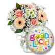 A fresh, uplifting gift set photographed against a clean white backdrop: a generous glass vase holds a loose, garden-style bouquet of soft peach and light pink roses, warm peachy-orange Gerbera daisies with deep brown centers, and clouds of tiny white baby's breath (Gypsophila). Small white and lime-green chrysanthemum florets nestle through the arrangement, their crisp petals catching the light beside sprays of deep green foliage that give the composition breathing space and natural depth. The vase is simple clear glass, the stems visible through the water, finished with a rustic twine bow that lends a hand-tied, artisanal feel. To the right, slightly overlapping the lower bouquet, sits a round metallic silver foil balloon printed with a cheerful Good Luck message in bouncy, multi-coloured letters. Two bright yellow smiley-face icons replace the o's in Good, one with a mischievous wink and the other with a playful tongue-out expression; scattered hearts, stars and colourful confetti motifs dance across the balloon, and a small Kaleidoscope logo is visible at its base. Soft studio light creates gentle highlights on the petals and a subtle sheen on the balloon, suggesting a quiet morning delivery meant to encourage and brighten. As a florist describing this piece, I imagine it handed over at the door before an important interview, exam or new chapter - a tactile bundle of scent, texture and colour designed to say I believe in you. Local Queensbury touches are in the thought behind it: easy to carry from the Broadway to a Jubilee line send-off, simple, reassuring and made to lift someone's spirits.
