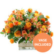 This image presents a joyful, harvest-toned bouquet of orange and yellow flowers, photographed sharply against a bright white background so that every nuance of colour and texture is clear, much like seeing a fresh delivery carried along Kingsbury Road on a crisp day. In the centre, generous orange roses open in layered spirals, their petals smooth and velvety, some showing a subtle gradient that shifts from fiery orange at the outer edge to a gentler, almost golden hue nearer the core. Around and between them, airy stems of peach and yellow alstroemeria add a lighter, more delicate feel; their narrow petals flare outward, each touched by fine stripes and speckles that offer a painterly detail. Punctuating the arrangement are clusters of small, round, vividly orange spiky blooms, reminiscent of safflower heads, their bristling texture standing in lively contrast to the softer roses and lilies. Encircling the whole display is an abundance of greenery: silvery eucalyptus leaves and deeper green foliage fan outwards, adding volume, depth, and a cool-toned frame that makes the warmer flowers glow even more brightly. The stems are gathered neatly into a clear, square-shaped glass vase, the transparency allowing a glimpse of the criss-crossed stems and the clean water that keeps them fresh, lending an honest, contemporary feel. Around the central band of the vase, a strip of natural burlap or jute is wrapped snugly, its woven, slightly rustic texture connecting the piece to homely, down-to-earth interiors, whether in a flat near Queensbury Station or a family home off Honeypot Lane. In the bottom right corner of the image, a strong orange circular badge announces VASE INCLUDED in white capital letters, clearly indicating that the container comes with the bouquet. The lighting is bright and even, with no distractions in the background, allowing the arrangement's cheerful character and crafted detail to shine, making it suitable for birthdays, thank-you gifts, or everyday cheer in Queensbury.