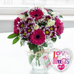 A richly textured bouquet and a whimsical balloon sit together in bright, gentle light, as if just delivered by a local Queensbury florist to mark a special moment. The flowers are arranged in a classic, rounded clear glass vase, their green stems fanning out neatly through the water. At the forefront, large gerbera daisies in deep fuchsia and magenta bloom with confident, open faces, their dark, velvety centres echoing the richness of the petals. Around them cluster vibrant chrysanthemums, each bloom a tiny marvel of colour with dark purple petals that fade to white at the tips and surround a sunny yellow core, adding contrast and a playful, almost confetti-like effect. Soft, creamy white roses peek out between these brighter flowers, their smooth petals layered in delicate spirals that speak of romance and calm. Light sprays of white baby's breath drift among the larger blossoms, like small tufts of cloud that make the arrangement feel airy and full, while flashes of fresh green foliage anchor the design and suggest a just-picked freshness. The vase is finished with a loop of rustic natural twine tied around its neck, from which hangs a small white tag bearing a pink rose symbol and green, handwritten-style lettering that adds a personal, handcrafted feel reminiscent of Flower Delivery Queensbury's bespoke creations. To the right, a shiny heart-shaped foil balloon, edged in silver, leans towards the blooms. Its white centre is printed with mosaic-style letters in shades of pink, purple, blue, and red that read Love n' Hugs 24-7, dotted with tiny hearts. Set on a pale, uncluttered surface against a softly blurred, bright background that suggests a sunlit Queensbury living room, this pairing radiates affection and is perfect for birthdays, romantic surprises, or simply sending hugs from afar.