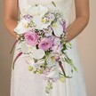 In a close-up scene set against a soft, neutral backdrop in Queensbury, a bride is shown holding an extravagant cascading bouquet that instantly captivates. The teardrop-shaped design overflows with white Phalaenopsis orchids, their ethereal petals highlighted by sunlit touches, supported by lush blush pink and lavender roses arranged skillfully at the bouquet's upper portion. Alongside, clusters of fluffy purple hydrangeas and interwoven sprigs of delicate baby's breath lend the arrangement a dreamy, cloud-like quality, while slender strands of rich magenta Amaranthus cascade alongside deep green foliage for dramatic depth. Subtle buds and wisps of lighter purple blooms create gentle movement and softness. The bride herself is seen from mid-torso to waist, wearing a white dress with detailed lace that echoes traditional local bridal styles, the soft drape of her skirt pooling below. Each element stands out vividly against the understated cream wall, ensuring that the artistry and play of colour-white, lavender, blush, and fresh greens-are the focus. This bouquet, prepared with the refined touch of Queensbury's florists, encapsulates both classic romance and fresh creativity, serving as inspiration for any couple wishing to blend elegance with a dash of local charm on their wedding day.