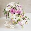 This image portrays an elegant, romantic floral centerpiece, the kind you might find gracing a wedding table at a small reception venue in Queensbury or a beautifully set dining room near Queensbury tube station. At the heart of the scene is a round white wicker basket, its woven pattern crisp and intricate, overflowing with carefully arranged blooms. Several luxurious pastel pink roses, fully open and spiralling out from tight centers, bring a sense of timeless romance. Surrounding them are serene white Phalaenopsis orchids, their petals smooth and slightly translucent, stretching out like butterfly wings around vibrant yellow and soft orange throats that add detail and sophistication. Ruffled white Lisianthus, some brushed with gentle purple along the petal edges, tuck in between the larger flowers, while pale pink stock stems climb upward and drift over the rim, lending height, texture, and a suggestion of sweet fragrance. Airy sprays of white baby's breath weave through the arrangement, softening transitions and creating a delicate, cloud-like veil around the focal blooms. Fresh green leaves and slender buds peek through at the edges, grounding the pastel palette with a touch of natural energy and movement. Resting on the bright white tabletop in front of the basket are two decorative woven spheres, formed from fine, pale strands of natural material, echoing the basket's weave and adding a sculptural, minimalist touch. Behind everything, a softly textured, neutral fabric background-somewhere between beige and light taupe-gently recedes, ensuring the colours of the flowers appear crisp yet calming. The overall impression is one of quiet luxury and heartfelt celebration, perfectly suited for a Queensbury wedding breakfast, anniversary meal, or special family gathering where fresh flowers play a central role in the table décor.