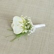 A delicately crafted white orchid boutonniere rests on a softly textured, light beige woven fabric, the kind of natural linen-like material you might see on a rustic table in a Queensbury townhouse near the high street. At the heart of the piece is a flawless Phalaenopsis orchid bloom, its broad, creamy white petals opening in a gentle curve. The petals have a subtle, satiny sheen, and in the centre you can just discern a whisper of pale yellow deep in the throat, lending a quiet warmth and life to the flower. Encircling this central orchid are fine, cloud-like clusters of white baby's breath, each tiny Gypsophila bloom creating a misty halo that softens the boutonniere's outline. Threaded in between are slender strands of feathery green foliage, likely a delicate fern, adding notes of fresh woodland green that contrast beautifully with the crisp whites. The stems are carefully aligned and wrapped in a pristine white satin ribbon, which is tied into a neat, understated bow near the base, suggesting the florist's precise hands and years of practice. Soft, even lighting falls across the scene, revealing the grain of the fabric background and the gentle shadows cast by the petals and sprigs. The overall mood is calm, elegant, and quietly romantic, evoking a wedding morning in Queensbury as a groom prepares for his ceremony at a local venue, perhaps near one of the small parks or community halls. This boutonniere feels ideal for a groom or groomsman seeking an accessory that speaks of purity, fresh beginnings, and timeless style, the kind of detail that completes a carefully chosen suit without overwhelming it.