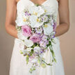 The image shows a waist-up view of a bride holding a cascading bridal bouquet that feels both opulent and serene, framed against a soft, neutral beige backdrop that lets the flowers take centre stage. The bouquet is composed around sweeping white Phalaenopsis orchids, their broad, droplet-shaped petals almost glowing, each blossom accented with a warm yellow-orange centre that adds a hint of tropical sophistication. Nestled closely among them are full-headed pink roses and dusty lavender-pink garden roses, their spiralled petals forming plush rosettes that suggest classic romance and careful craftsmanship. Tiny clusters of white lisianthus with faint purple tinges at the petal edges echo the pastel theme, while cloud-like gypsophila drifts between the larger blooms, lending an airy, dreamlike texture. Slim spires of pale lavender stock introduce a subtle vertical rhythm and a soft, fresh fragrance that one can almost imagine rising from the bouquet. The arrangement flows naturally downward in a teardrop silhouette, with flowers and delicate greenery tapering towards the hem of the dress, creating a sense of gentle movement as if it could sway with every step down the aisle. The bridal gown itself is simple and strapless, its clean white fabric providing a calm backdrop that mirrors the bouquet's purity. As a Queensbury florist, I can picture this design being carried into intimate ceremonies near the quiet residential avenues off Honeypot Lane, or photographed in the soft light around local gardens. The photograph beautifully conveys a refined wedding style, ideal for couples seeking cascading bouquets, pastel wedding flowers, and bespoke designs available for delivery throughout Queensbury.