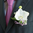 Pinned neatly to a dark, sharply pressed suit jacket, this orchid boutonniere carries the calm, polished atmosphere of a special day unfolding somewhere in Queensbury, perhaps just after an intimate ceremony. The focal point is a single white orchid, its petals full and velvety, opening in a poised, symmetrical curve. Their surface catches the light with a soft, porcelain-like glow, and at the edges there is the faintest whisper of ivory, adding depth to the pure white. Surrounding the orchid, cool-toned foliage in silvery and light green tones nestles close, their leaves slightly textured, offering a natural contrast to the smoothness of the bloom. Sprigs of lilac flowers and tiny white buds weave delicately through the greenery, adding touches of gentle colour and a soft, almost powdery fragrance that recalls a garden border along a quiet Queensbury street. The stems are bound in a slim violet ribbon, tied with deliberate precision so it lies flat and elegant, its satin finish reflecting the subtle shimmer of the purple-lilac tie beneath the lapel. Together, flower, foliage, and ribbon create a composition that looks balanced and intentional rather than showy, as though the wearer has chosen one perfect detail rather than many. The boutonniere rests just above the pocket, creating a strong contrast against the charcoal fabric of the suit, so the orchid appears almost to float. There is a mood of anticipation here-of toasts waiting at a nearby reception venue, photographs taken against local brickwork and greenery, and a quiet happiness captured in one carefully crafted floral piece.