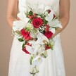 A radiant bridal bouquet is captured in a detailed, medium-close shot, held gently by a bride in a flowing white gown, her arms bare and posture serene. The bouquet is designed in a dramatic teardrop cascade, with brilliant white lilies-large and trumpet-shaped-standing out among a harmonious medley of crimson roses. Soft phalaenopsis orchids trail at the base, lending movement and delicate intricacy to the arrangement. Accentuating these main blooms are sprigs of white gypsophila, tiny blossoms woven between the layers for a touch of airy elegance, while clusters of red hypericum berries provide festive bursts of colour and textural contrast. The foliage includes a mixture of wispy, fine asparagus fern and structured green leaves, uniting the composition in fresh, local character reminiscent of gardens near Queensbury Station. Set against a light neutral background, the bouquet's colors and forms are fully showcased, creating a refined, romantic vision perfect for bridal parties or wedding photo shoots in Queensbury. Ideal for articles highlighting wedding floral trends, bouquet inspiration, or advice for local couples seeking timeless elegance, this arrangement embodies the exquisite craftsmanship and flair found at Queensbury's finest florists.