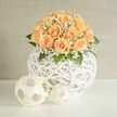 This arrangement reads like a study in tender colour and sculptural light: domed clusters of honeyed peach roses nestle tightly together, each bloom showing a subtle gradient from pale cream to ripe apricot, while delicate white gypsophila threads through the composition like a soft veil. The flowers rest upon a striking spherical basket woven in white rattan; its open lattice allows light to pass and create a floating sensation, so the bouquet looks both modern and weightless. As you imagine running a thumb along a petal you would note the velvety surface and the faint, sweet perfume that roses carry-complemented by the powdery freshness of the baby's breath and the cool, green counterpoint of foliage tucked beneath. The textured basket gives the display an architectural base: the criss-cross lines echo the blooms' spirals and create a pleasing interplay of shadow and highlight. This piece suits a gentle celebration-an intimate wedding, a milestone quietly observed, or a tasteful gift to mark new beginnings-and is crafted with the care local clients expect near Queensbury station, serving homes and small venues in Queensbury, Kingsbury and Wembley. At Flower Delivery Queensbury we arrange each stem deliberately, aiming for an effect that reads both close up and across a room: calm, refined, and full of feeling without excess.