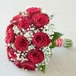A radiant hand-tied bouquet rests upon a clean, pale backdrop-a true Queensbury classic brought to life by the skilled hands of a seasoned local florist. The rounded cluster of over a dozen deep red roses forms the heart of the design, each rose at the height of bloom, displaying satiny, intricate petals that invite one's gaze. Interlaced throughout are airy white baby's breath blossoms, their countless tiny florets lightening the ensemble and adding a visual softness reminiscent of morning mists over nearby local parks. The occasional dark green leaf peeks through the mass of florals, providing an organic accent that mirrors the leafy avenues of Kingsbury Road. The handle, expertly wrapped in wide, vibrant red satin ribbon with crisp white stripes and a subtle spiral of tiny pearls, imparts an added air of delicacy and romance. Beneath, neatly trimmed stems emerge-fresh, green, and meticulously aligned-signifying the bouquet's readiness for any special event. The entire composition glows under even ambient light, highlighting every layered petal and sparkling embellishment. Rich with symbolism-red roses for ardent love, baby's breath for purity and innocence-this arrangement embodies timeless romantic gestures. Perfect for modern weddings in the Queensbury area, anniversaries, or as a grand romantic surprise, each element is selected to reflect both tradition and local charm, capturing the essence of celebration in every detail.