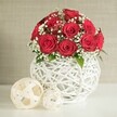 An elegant cluster of deep red roses stands out vividly against a calm, neutral backdrop, creating a romantic table arrangement that would look at home at any intimate Queensbury wedding. The roses, around a dozen to fifteen in total, are gathered into a neat, spherical bouquet, each flower fully open with densely layered petals that appear soft and velvety to the touch. Their intense crimson hue conveys passion and love, while light, airy sprigs of white baby's breath weave among the blooms, lending a lace-like softness reminiscent of bridal veils seen at local ceremony venues near Queensbury. A few discreet green leaves punctuate the composition, adding gentle touches of natural colour without disturbing the refined balance. The bouquet is presented in a distinctive round white vase made from interlaced strands that form a hollow, openwork design, similar to a carefully woven basket with a modern twist. This sculptural vase allows light to filter through its gaps, casting delicate shadows on the smooth white tabletop beneath and enhancing the sense of depth and craftsmanship. To the left of the vase sit two decorative woven balls in warm off-white tones: the larger sphere occupies the foreground, while the smaller one sits slightly behind, their open lattice patterns echoing the vase and tying the whole scene together. The background is a softly textured, pale wall, intentionally understated so the vivid red roses and intricate textures can command full attention. The composition suggests quiet celebration and thoughtful design, ideal for wedding receptions, anniversary dinners, or sophisticated home decor across Queensbury and neighbouring areas.