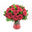 A lavish bouquet of deep crimson red roses stands proudly in a ruby-red glass vase, its rich tones glowing as if lit from within. Each rose is tightly clustered with its neighbours, creating a generous dome of velvety blooms that seem almost to spill over the rim. The petals form intricate spirals, some still wound in tight, secretive centers, others opening into full, cup-shaped flowers that reveal subtle variations of scarlet and ruby at their edges. Around and between the roses, lush dark green foliage, likely Ruscus or a similar filler green, fans out in polished, tapered leaves, adding depth and a cool contrast to the warmth of the red. The greens feel fresh and supple, as if they were just conditioned in a busy Queensbury flower studio before being arranged with care. The vase itself is a translucent ruby-red glass with an elegant twisted or spiralled pattern etched along its surface, catching the light in slender, shimmering ribbons. Its gently flared top and solid base lend a sense of balance and classic formality, while the stems, visible through the coloured glass, add a subtle sense of structure. Set against a pristine white background, the bouquet becomes the sole focus, its colours intensified by the clean, uncluttered space around it. The even, bright lighting highlights every petal and leaf, evoking the feeling of freshly delivered roses ready to grace a dining table in a Queensbury townhouse, a romantic corner near Queensbury tube, or a quiet celebration at home. The arrangement suggests love, devotion, and timeless romance, perfect for anniversaries, Valentine's surprises, or heartfelt gestures of admiration shared across the local community.
