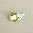 A single white rose boutonniere lies diagonally across a soft, linen-like beige fabric, its creamy petals just beginning to unfurl as if caught at the very first moment of bloom. The rosebud is a gentle ivory rather than stark white, with a faint hint of green at the base where the sepals cradle the flower, giving a sense of freshness as though it has just been prepared in a Queensbury florist's workshop before a wedding at the local registry office. Around the rose, tiny tufts of white baby's breath cluster like little clouds, each minute blossom forming a light halo that softens the overall shape and adds a romantic, almost misty texture. Slender sprigs of deep, glossy green foliage and finer, fern-like leaves frame the bloom, their lines leading the eye along the boutonniere and creating a subtle contrast against the pale backdrop. All of the stems are neatly bound together in a smooth white satin ribbon, wrapped tight and finished with a small, perfectly tied bow that sits just beneath the flower head, suggesting careful, practiced hands have worked each fold. The fabric beneath has a visible diagonal weave, reminiscent of natural linen often used on tables in Queensbury's intimate wedding venues, its understated pattern highlighting rather than competing with the flowers. Soft, diffused light falls from one side, casting delicate shadows that emphasise the gentle curves of each petal and the airy spaces between the baby's breath. The overall mood is calm, pure, and quietly celebratory, the kind of classic rose boutonniere a groom or groomsman might wear to express timeless elegance and new beginnings during a wedding or formal event in and around Queensbury.