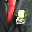 This close-up image lingers on a classic wedding boutonniere, captured as if just before the ceremony begins in a quiet corner of Queensbury. A single ivory-white rose forms the centrepiece, its petals gently unfurling in smooth, velvety layers that rise from a creamy heart to soft, rounded outer edges. The bloom sits proudly against a richly textured charcoal lapel, the contrast giving it a luminous quality, like a small beacon of calm in the anticipation of the day. Around the rose, clusters of snowy baby's breath nestle close, each tiny flower no larger than a pinhead, adding a cloudlike halo of softness and innocence. Threaded between them, slender loops of polished greenery and fine fern sprigs arc gracefully, introducing a fresh, woodland feel that never competes with the rose's quiet authority. The stem is bound tightly and finished with a neat white satin ribbon, the bow carefully centred, its ends cut with precision, suggesting the steady hand of an experienced florist who has dressed many grooms and prom guests across Queensbury, Kenton, and the surrounding streets. The overall mood is refined and ceremonial, the kind of detail chosen for weddings in local venues or formal celebrations that matter deeply to families. Though the image is still, you can almost sense the rustle of the suit fabric and the soft scent of the rose, clean and unobtrusive. This boutonniere expresses tenderness and tradition in equal measure, an understated flourish that quietly tells everyone present: this is a day where love, commitment, and shared memories take centre stage.