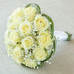 There is a dreamlike calm to this classic wedding bouquet, photographed from just above to show the perfect domed silhouette of cream roses and baby's breath. Around fifteen to twenty creamy yellow-ivory roses sit closely packed, their petals curling inward to soft spiral centres that catch a pale, diffused light and reveal subtle tonal shifts from ivory to warm vanilla. Sprays of delicate Gypsophila weave between the larger blooms, creating a feathery veil that softens the composition and lets the roses breathe. Curving strands of bright green lily or bear grass trace a gentle heart-like outline around the bouquet, adding a modern line without stealing the vintage romance. The stems are visible at the bottom, bound neatly in smooth white satin ribbon that lies cool and compact in the hand-a detail brides appreciate as they walk down the aisle. The bouquet rests on a lightly textured taupe-linen surface that amplifies the softness of the palette and suggests a quiet bridal suite bathed in morning light. The aroma is subtle: the honeyed sweetness of cream roses balanced by grassy freshness, a scent that carries well through vows and photos. As an experienced florist serving Queensbury, and used frequently by couples planning ceremonies across Queensbury, Kingsbury and Harrow, I craft each stem with attention to balance and longevity so the blooms travel well. This arrangement is a quiet statement of elegant restraint-perfect for ceremonies that favour timeless charm and sincere sentiment.